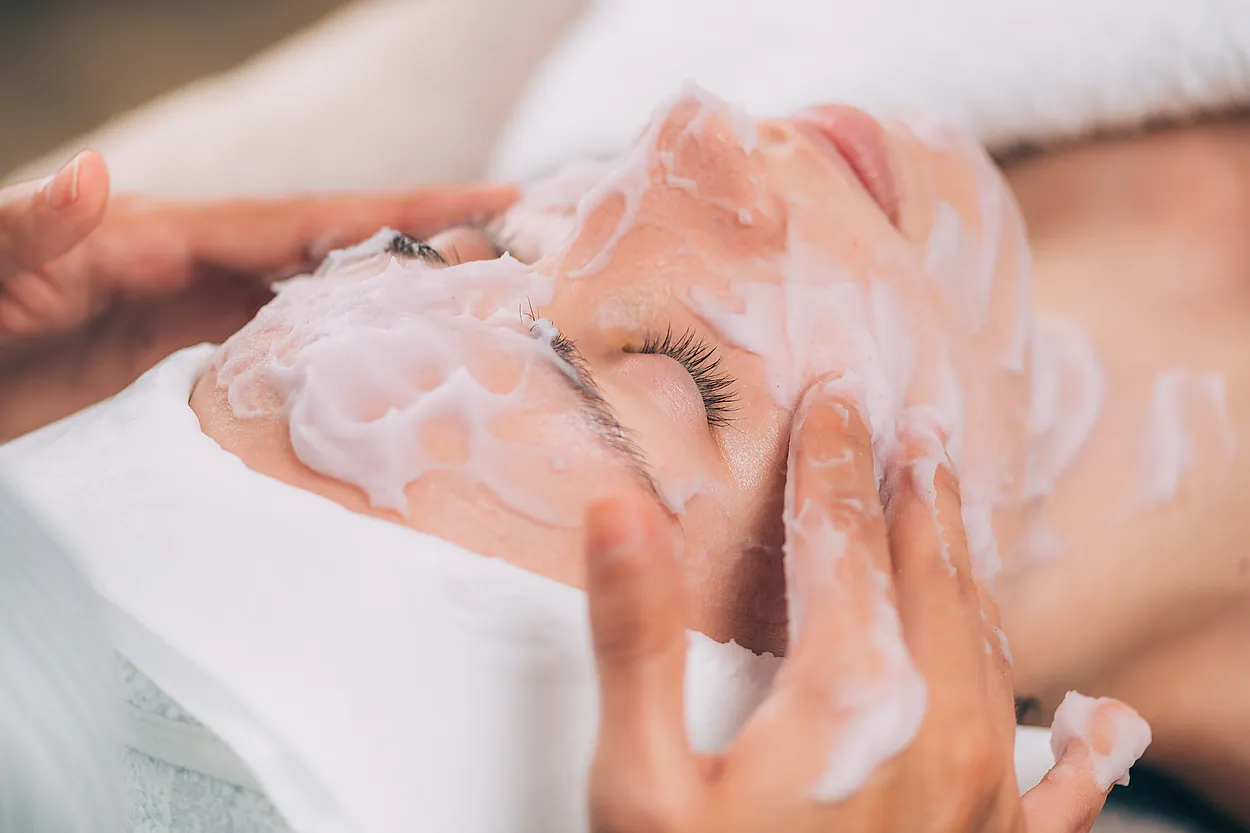 recorte manos de fisio untando crema en rostro de joven tumbada